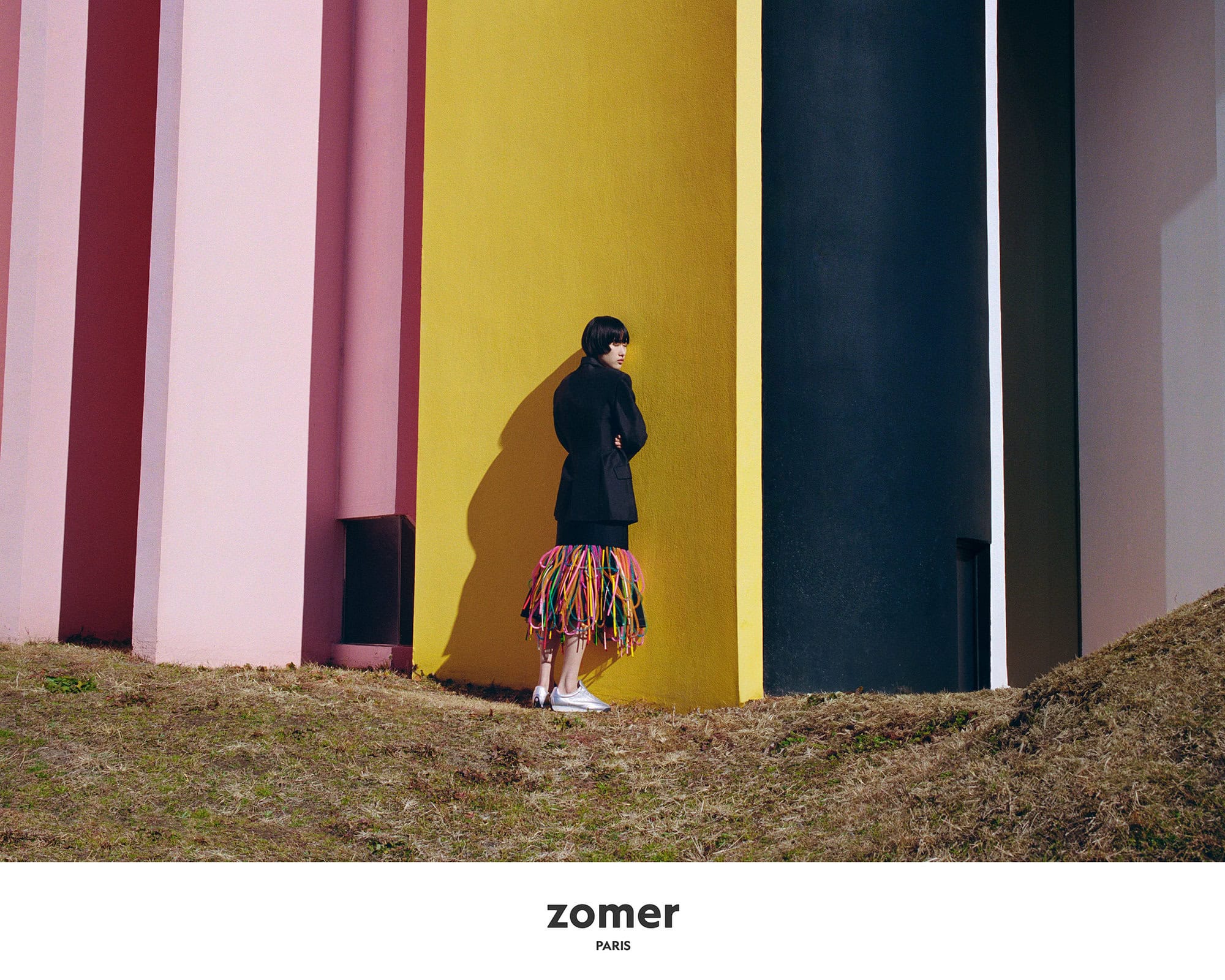 Model standing against a vibrant yellow and pink wall wearing a dark jacket and colourful fringed skirt outdoors in Japan.