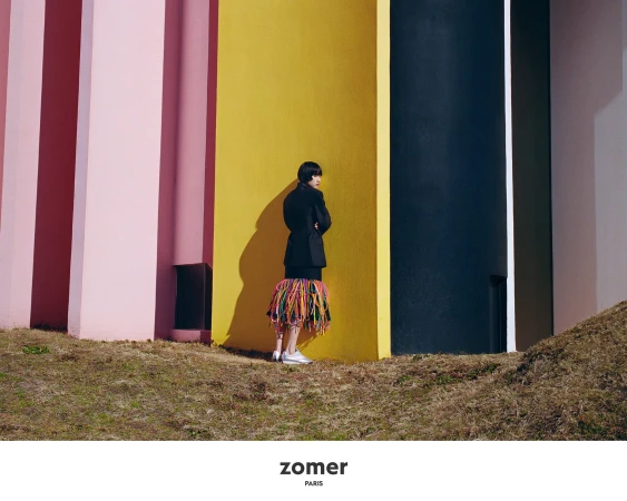 Model standing against a vibrant yellow and pink wall wearing a dark jacket and colourful fringed skirt outdoors in Japan.