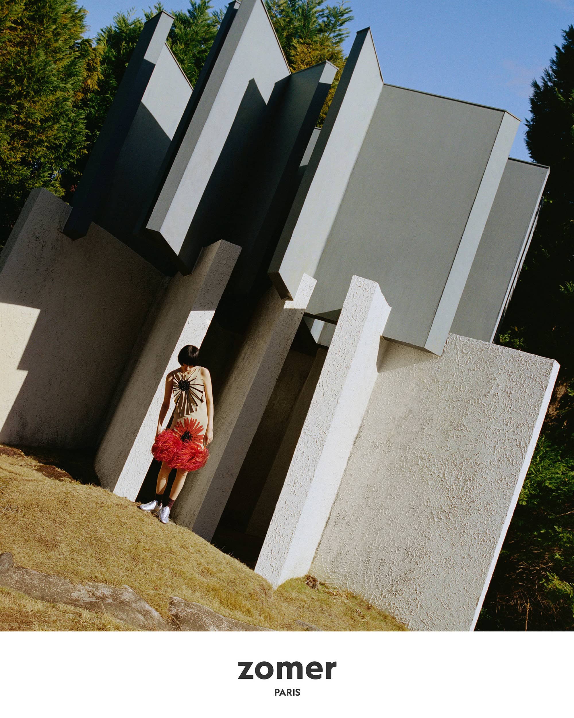 Model standing beneath a modern architectural structure wearing a textured dress with a red hem for Zomer spring ad campaign.