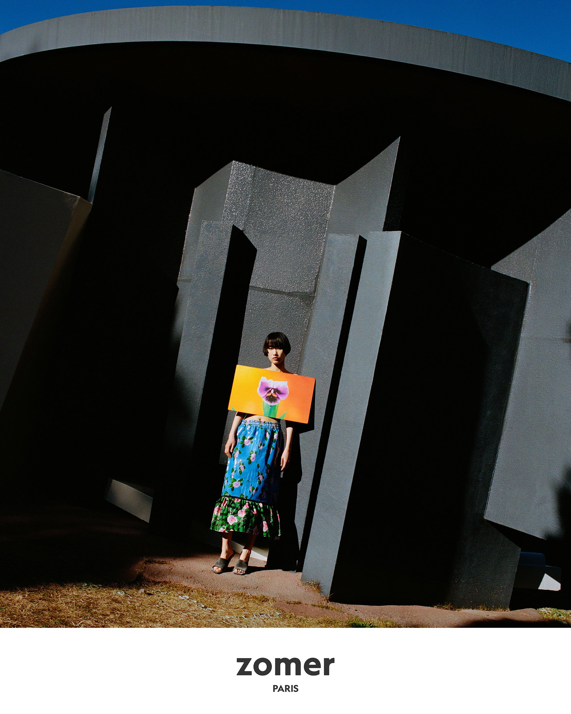 Model standing beneath a dark sculptural architectural structure holding a vibrant rectangular graphic panel