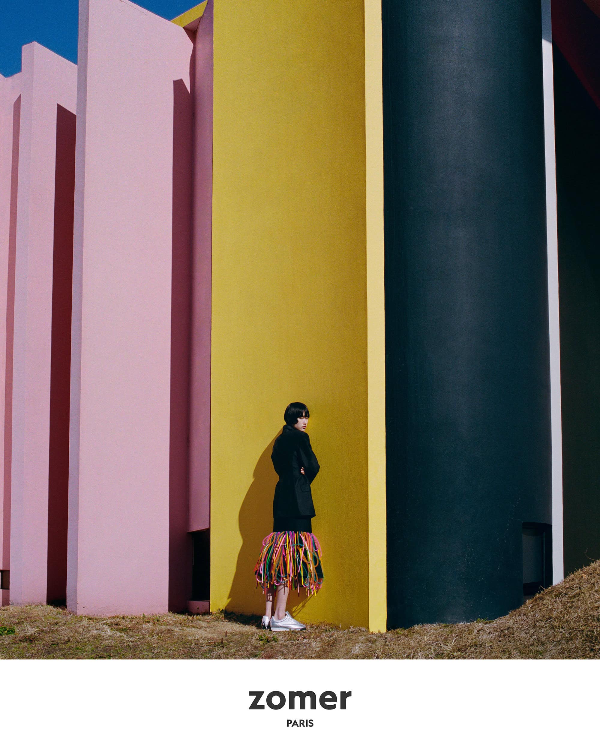 Model standing against bold yellow and pink architectural pillars wearing black coat with fringed hem