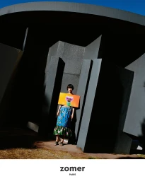 Model standing beneath a dark sculptural architectural structure holding a vibrant rectangular graphic panel
