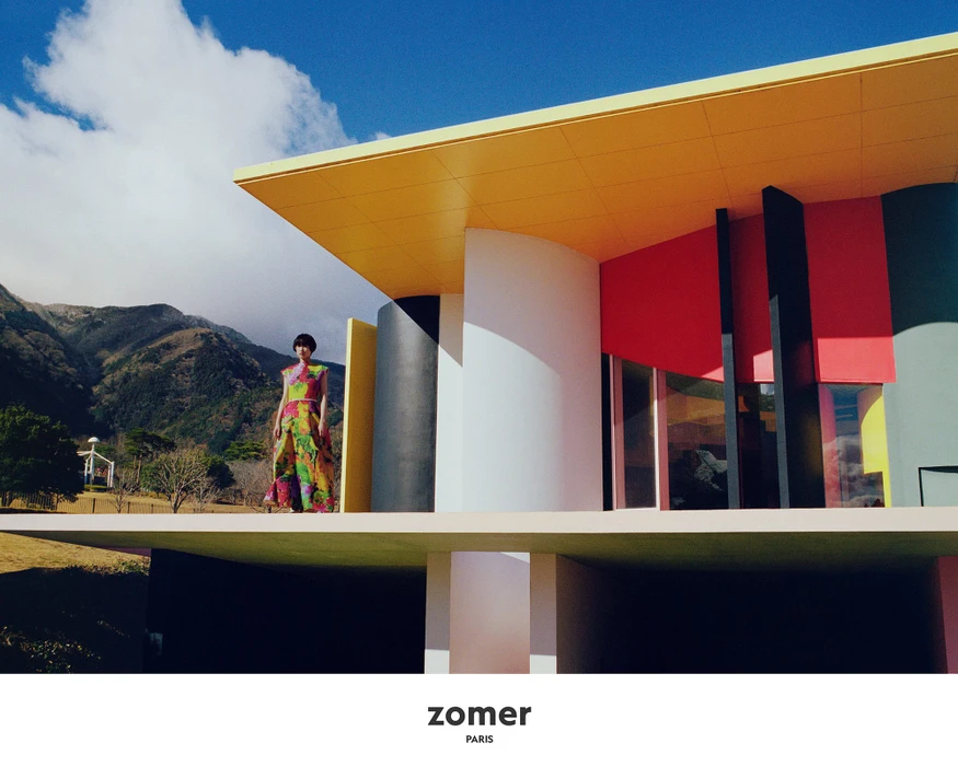 Model standing on a balcony of a brightly colored modern architectural building with mountains in the background