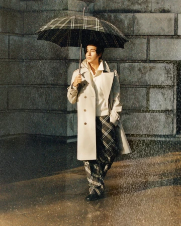 Man wearing a beige trench coat and plaid trousers holding a plaid umbrella outdoors in the rain against a stone building.