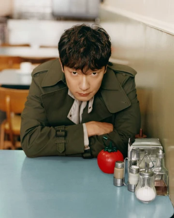Son Sukku wearing an olive green trench coat seated at a blue diner table for Burberry