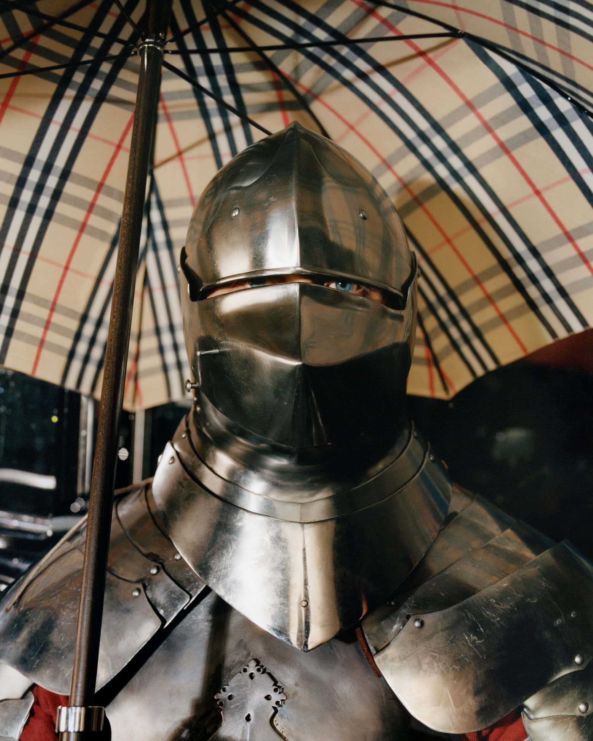 Close-up portrait of a person in polished medieval steel armor holding a Burberry check umbrella in London.