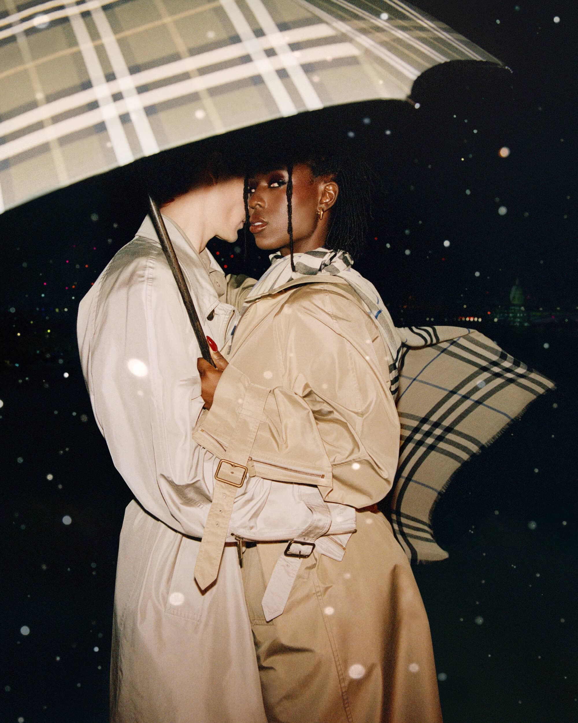 Two models wearing beige trench coats under a checked umbrella in the rain for Burberry by Drew Vickers.