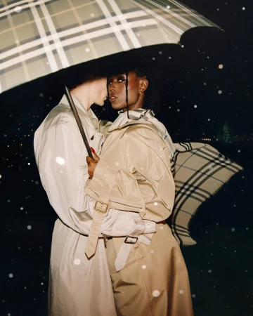 Two models wearing beige trench coats under a checked umbrella in the rain for Burberry by Drew Vickers.