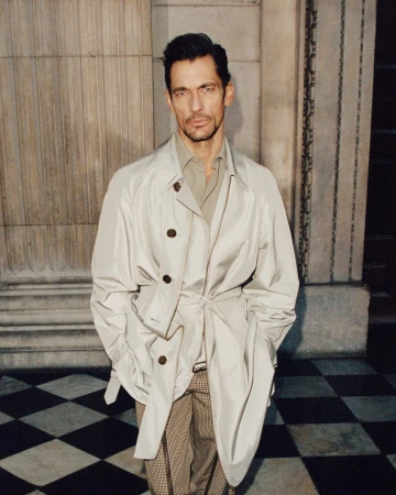 David Gandy wearing an off-white trench coat and patterned trousers standing against classic architectural stone columns.