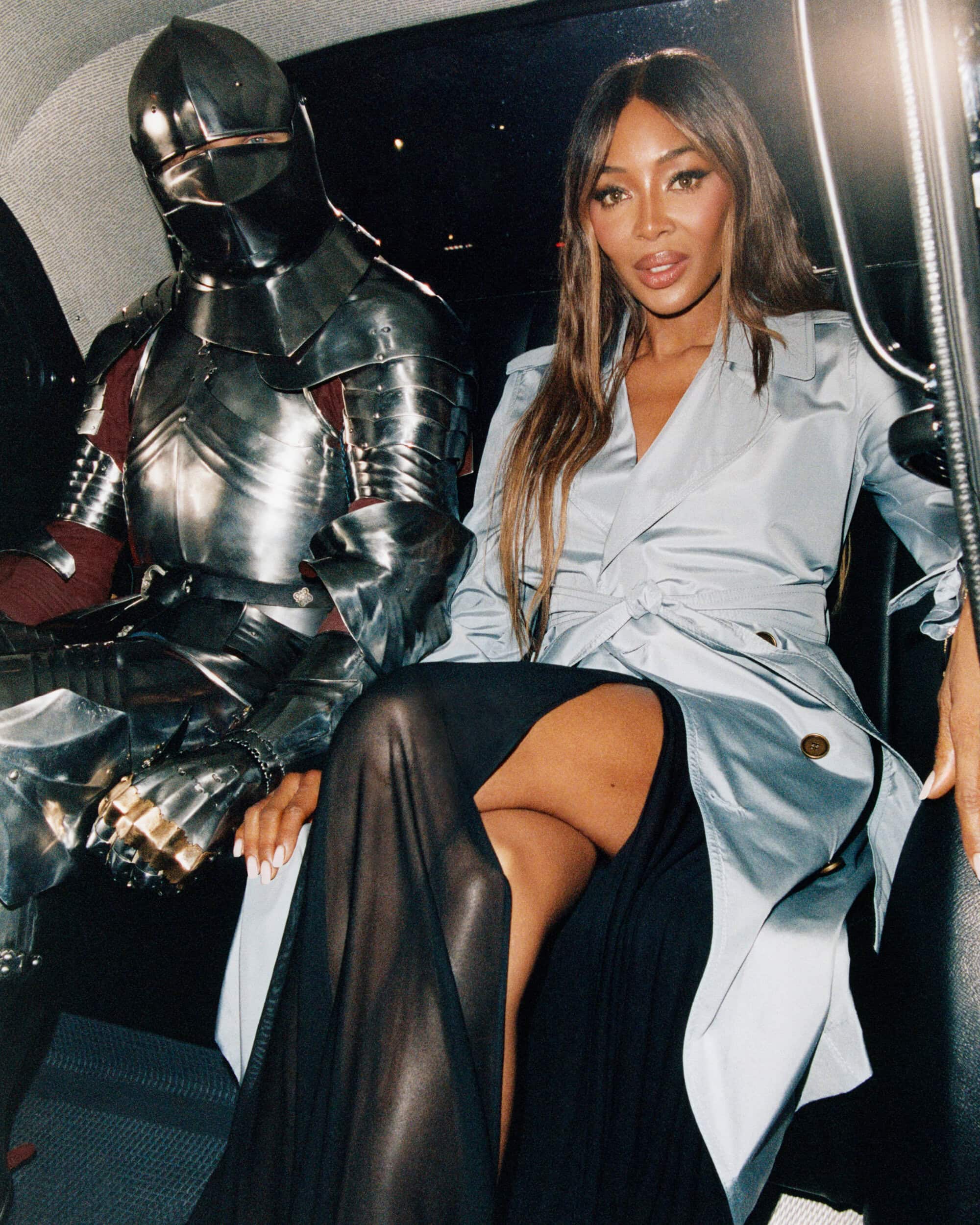 Naomi Campbell seated inside a vehicle next to a person in full metal suit of armor wearing a belted trench coat.