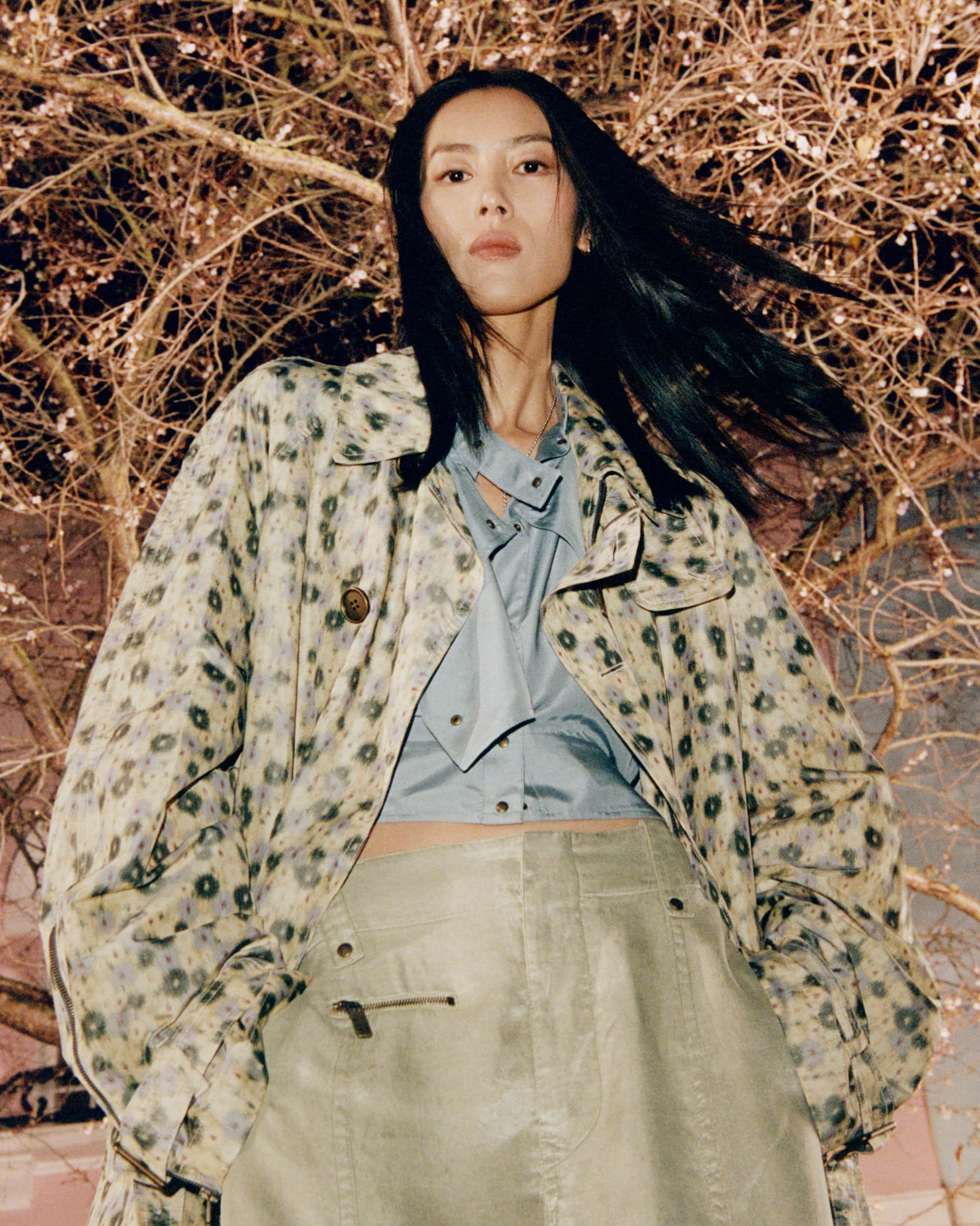 Model Liu Wen in patterned trench coat and blue shirt against bare branches in London for Burberry 2025 campaign.