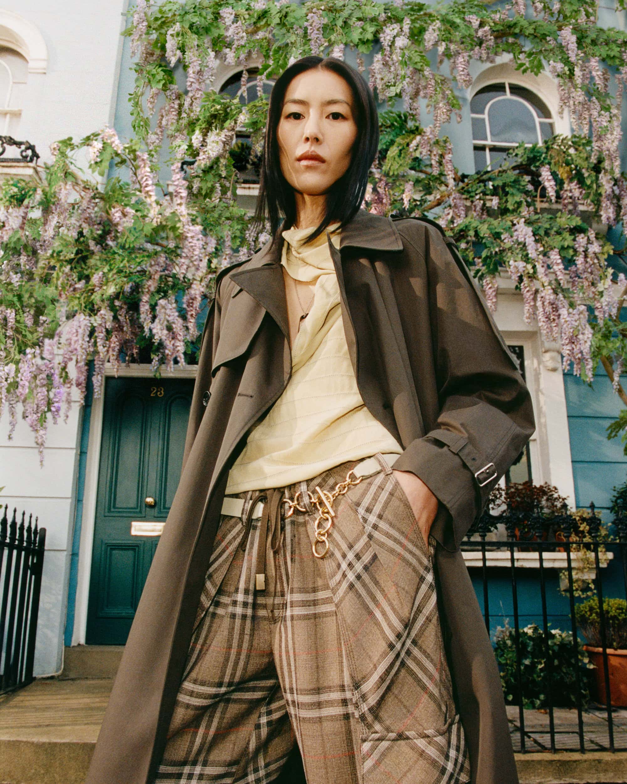 Model Liu Wen in a brown trench coat and plaid trousers posing in front of a wisteria-covered London townhouse
