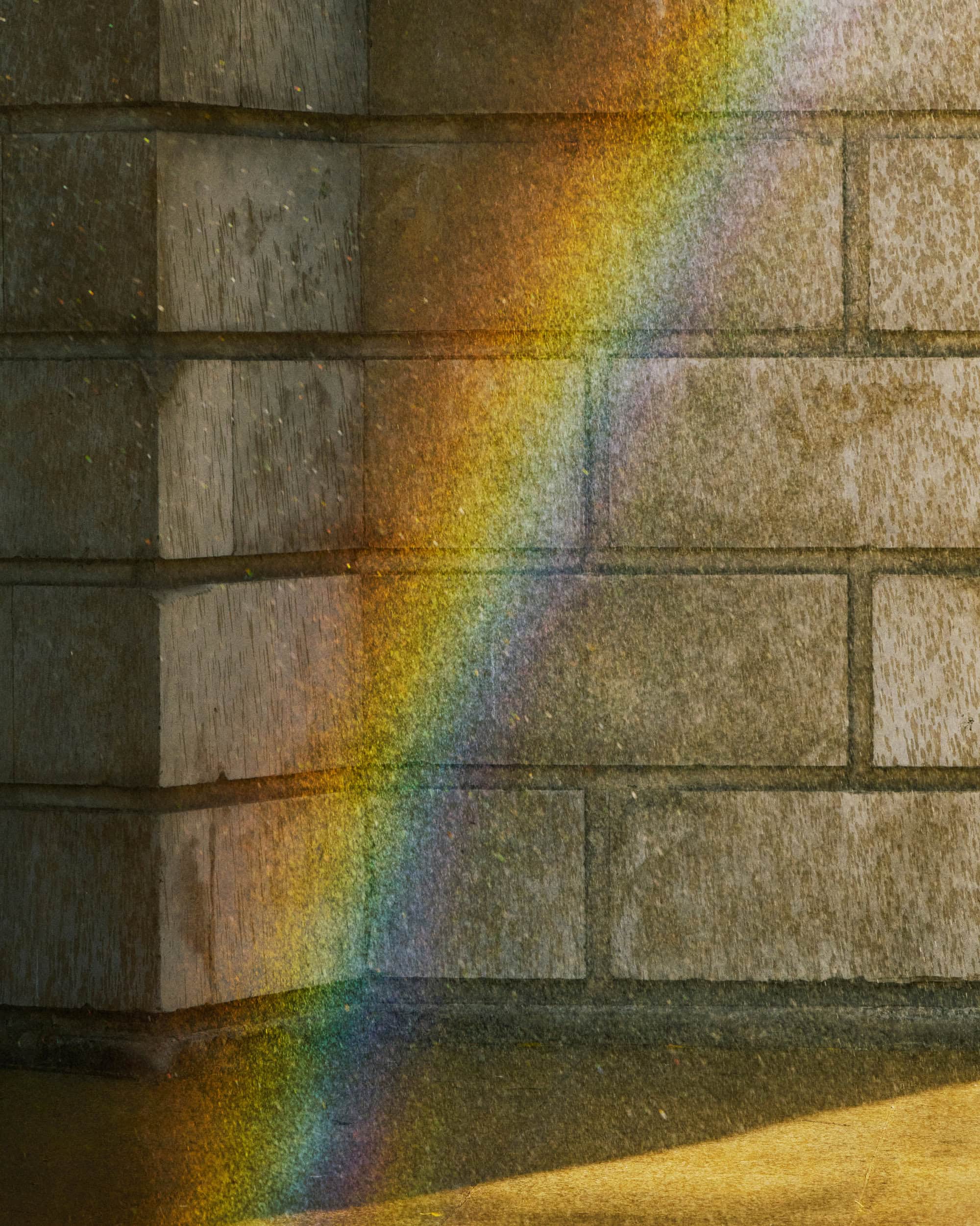 A diagonal beam of colorful light strikes a weathered stone wall corner, casting a vibrant prismatic spectrum across textured blocks.