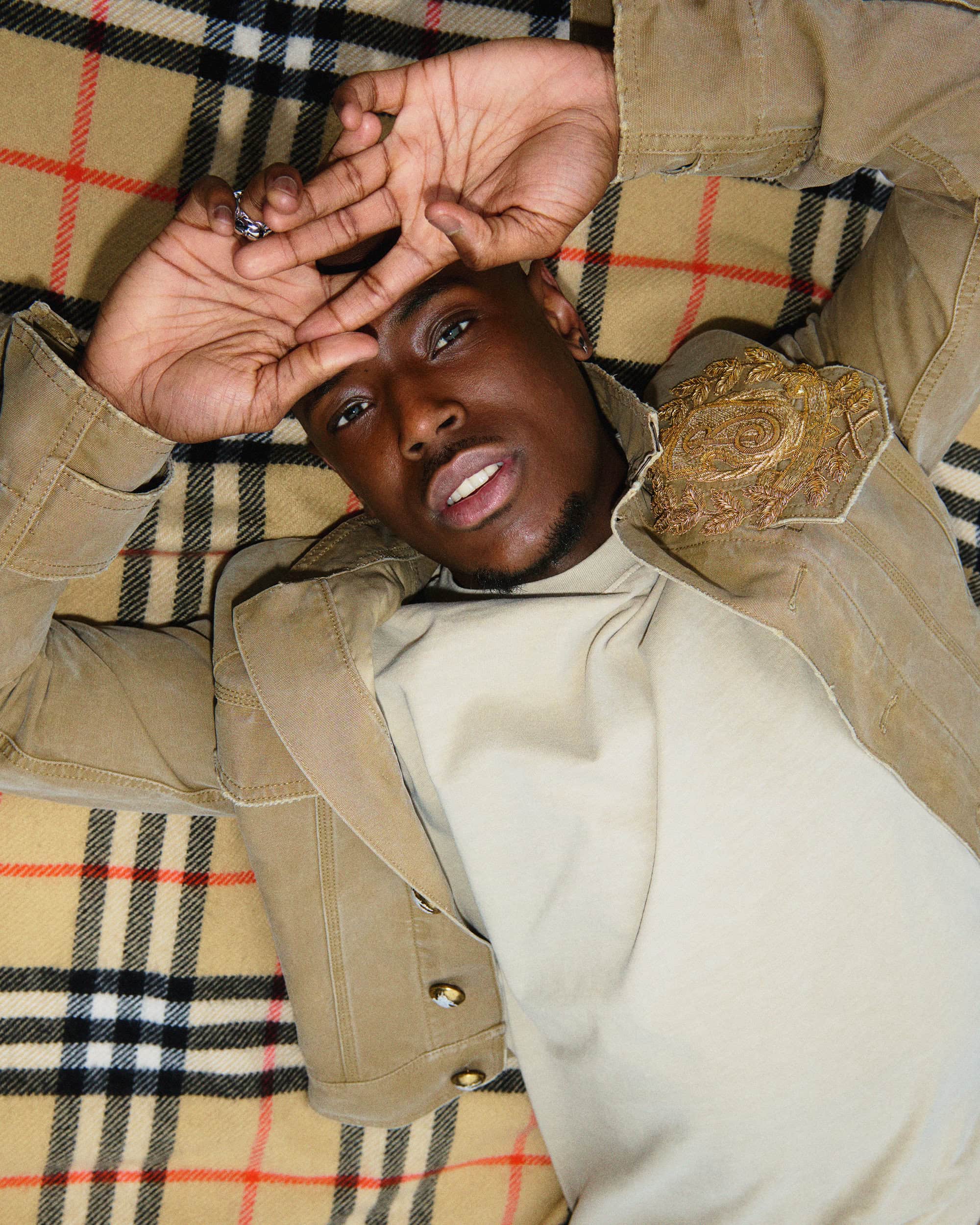 Close-up portrait of a Black man lying back on a beige and black plaid blanket wearing a tan jacket.