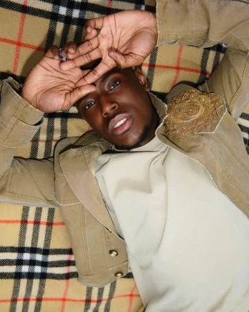 Close-up portrait of a Black man lying back on a beige and black plaid blanket wearing a tan jacket.