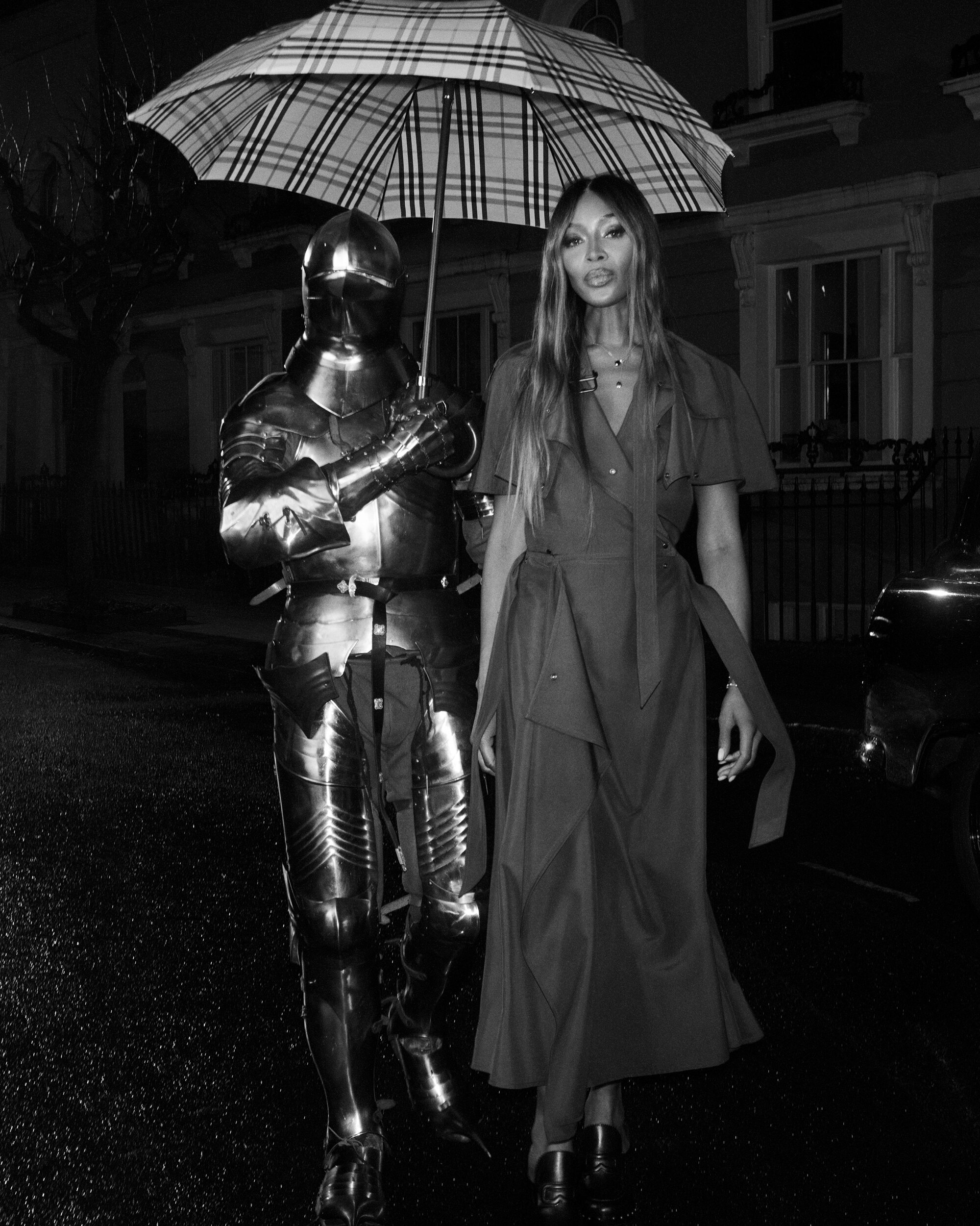 Naomi Campbell walking in London under a Burberry check umbrella next to a figure in full metal armor at night.