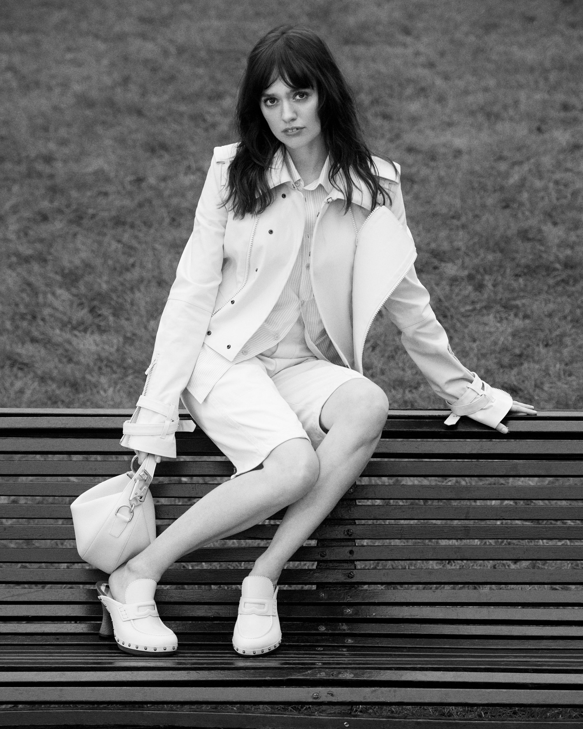 Woman in light-colored jacket and shorts seated on a park bench holding a handbag for Burberry ad campaign.