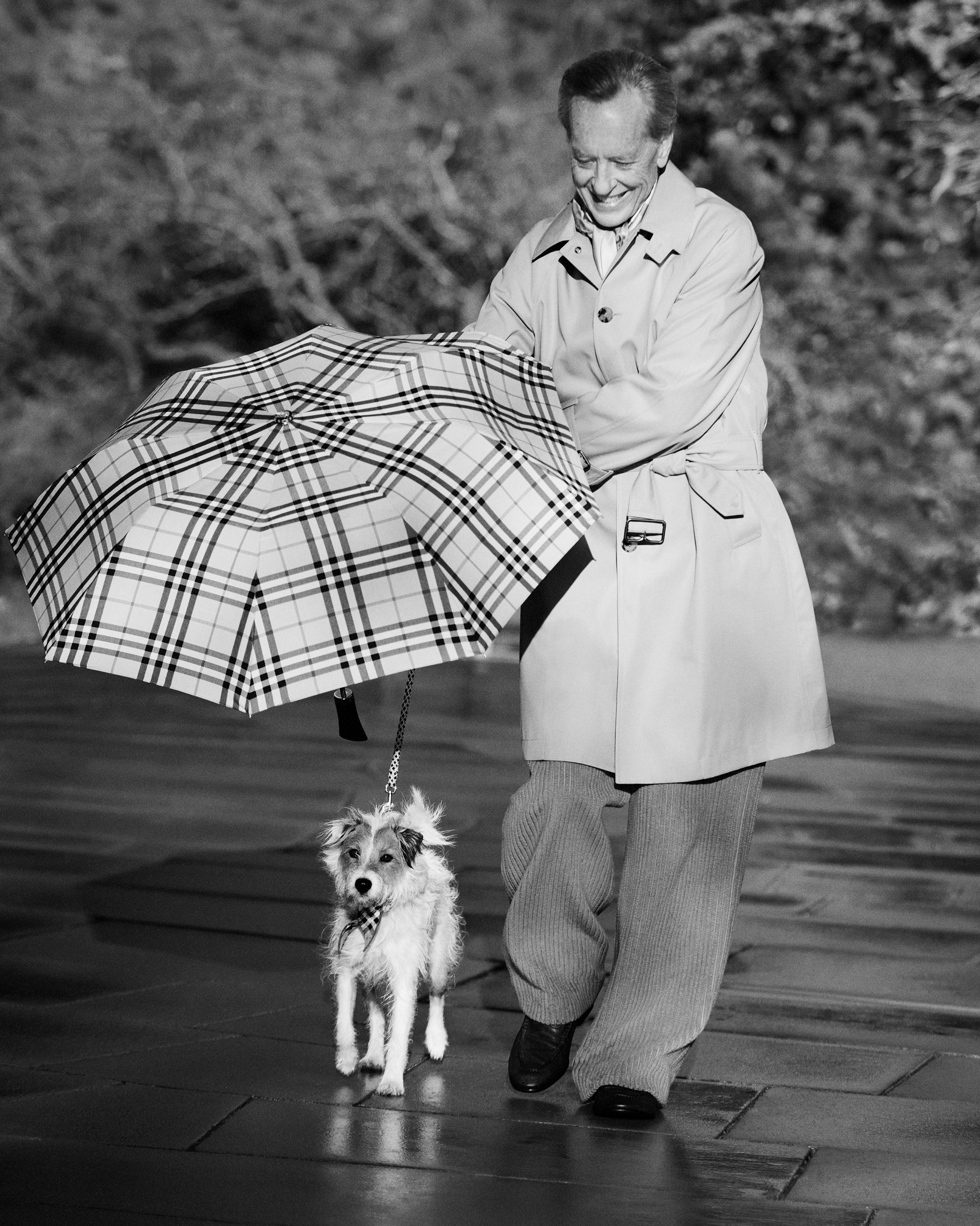 Richard E. Grant wearing a tan trench coat holding a plaid umbrella and walking a small dog on a paved path.
