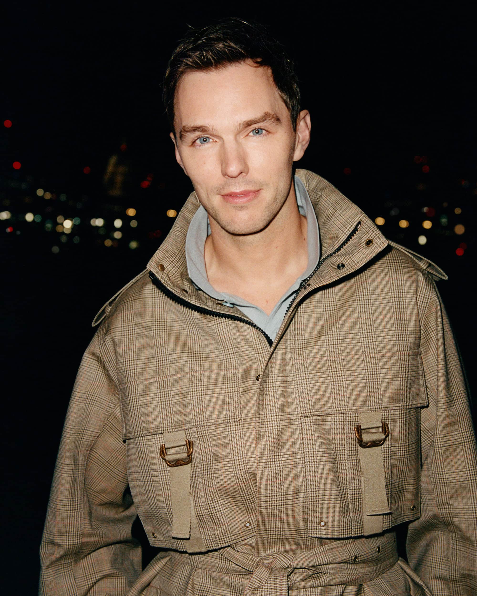 Portrait of Nicholas Hoult wearing a plaid belted jacket against a dark nighttime background with distant lights.