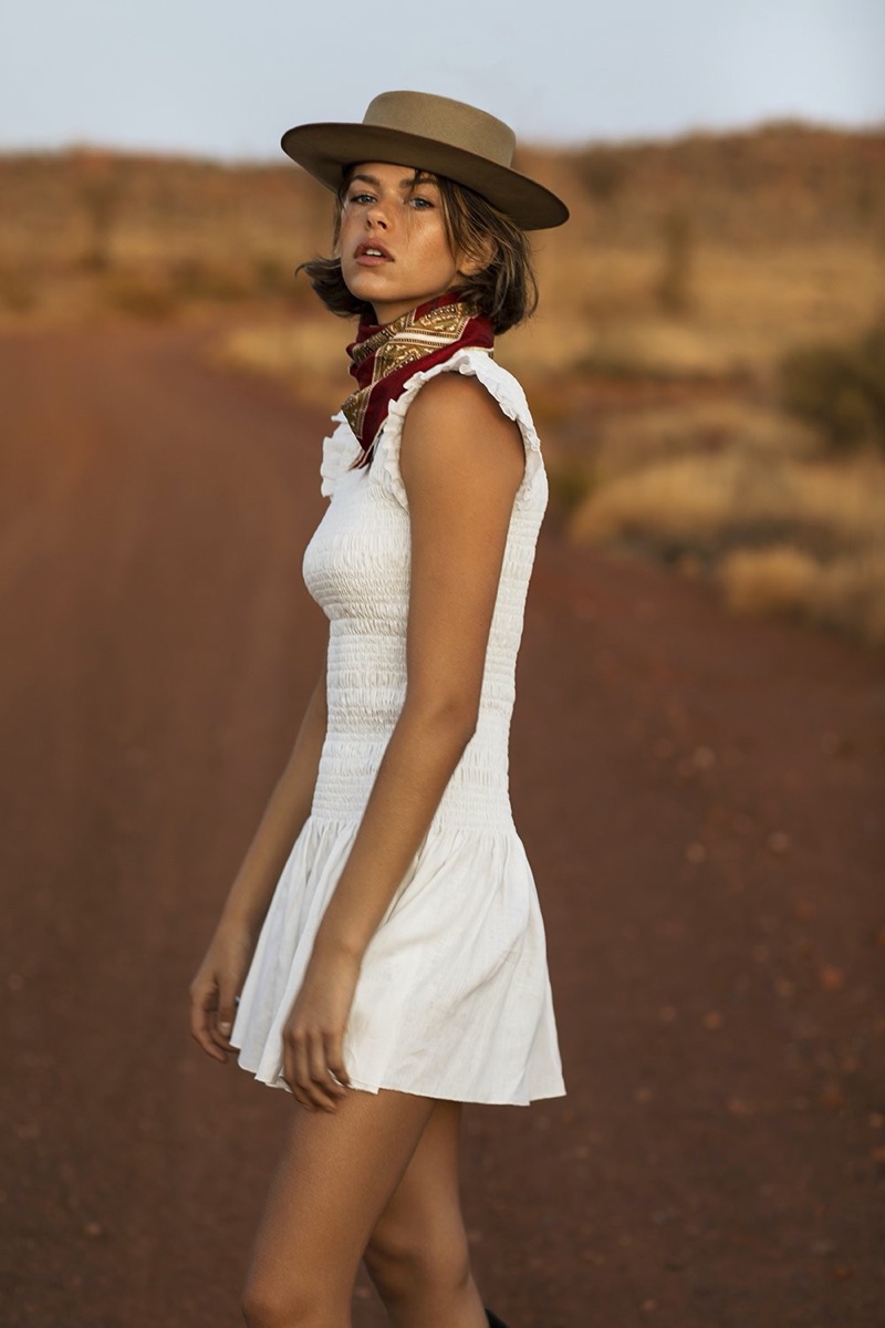 Woman in white dress and hat standing on a rural road with reddish-brown soil, wearing a patterned kerchief.