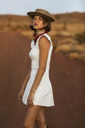 Woman in white dress and hat standing on a rural road with reddish-brown soil, wearing a patterned kerchief.
