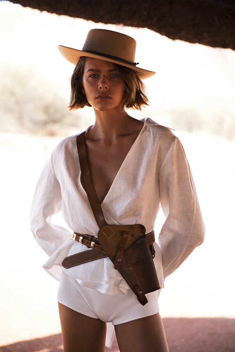 Woman with hat and holster stands outdoors in white blouse and shorts, creating a desert-like fashion editorial look with soft lighting.
