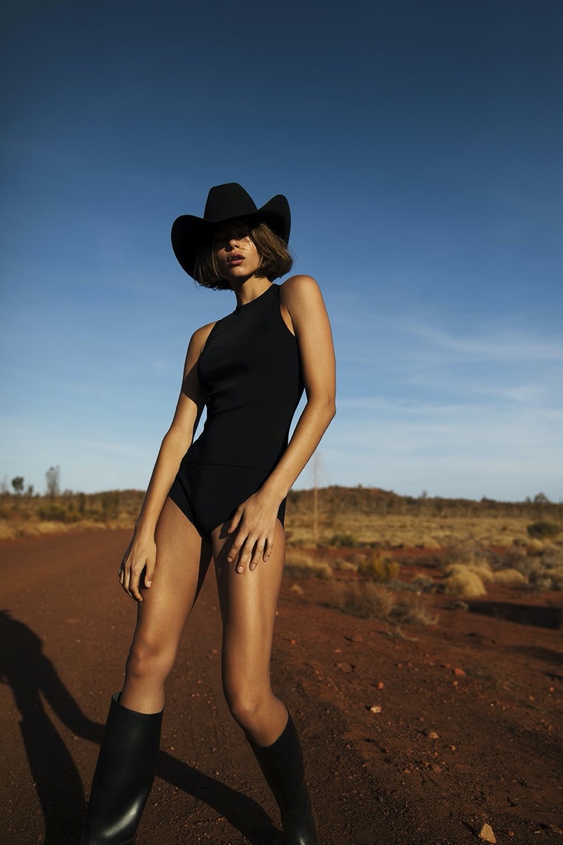 Adult female in black bodysuit and cowboy hat poses on red dirt road under blue sky in landscape shot, wearing knee-high black boots.