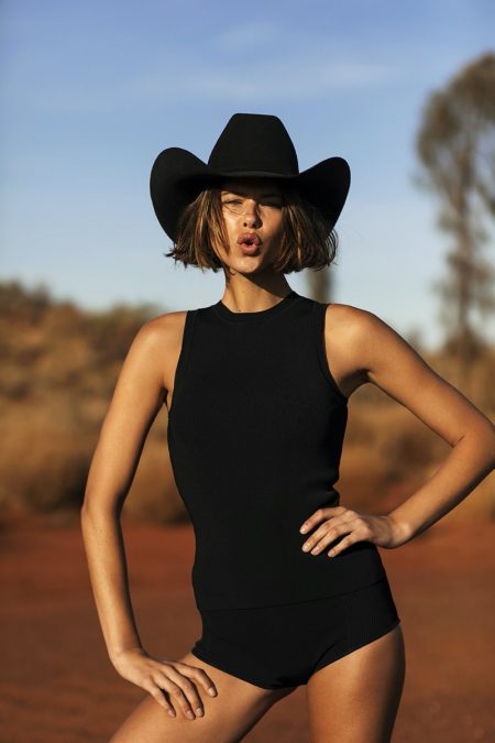 A woman poses in a black top, shorts, and cowboy hat against a desert backdrop, creating a striking fashion-editorial image outdoors.