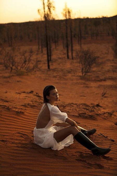 A woman in a white dress sits on a red sand dune in a desert landscape with scattered trees and a warm, orange sunset sky.