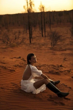 A woman in a white dress sits on a red sand dune in a desert landscape with scattered trees and a warm, orange sunset sky.