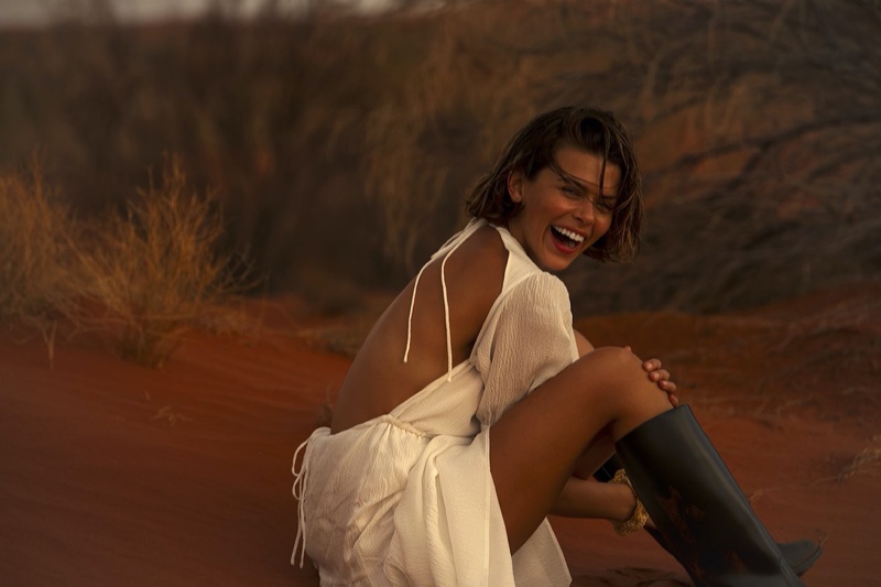 A smiling adult woman in a white dress poses in a sandy, outdoor environment. She is wearing tall, black boots and facing the camera.