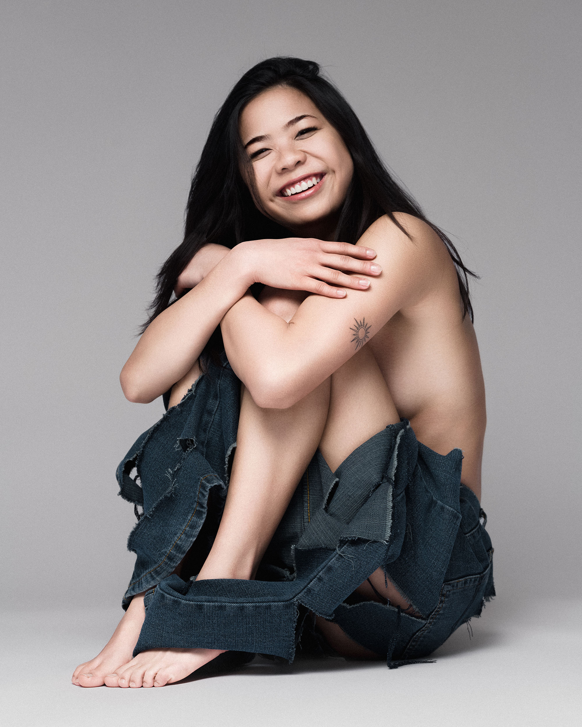 Young woman with dark hair smiles while seated, posing with an assortment of blue denim textile pieces in a neutral studio setting.