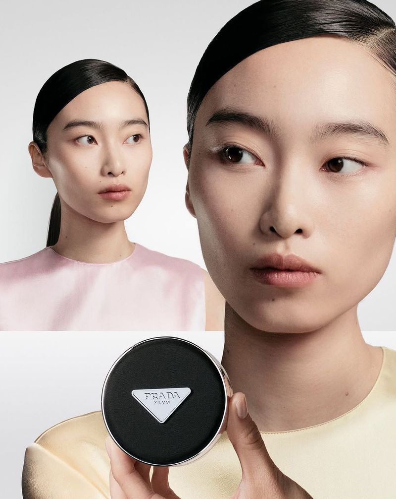 Cosmetic product shot featuring two adult Asian female models with smooth skin, holding Prada makeup compact with logo, clean beauty aesthetic.