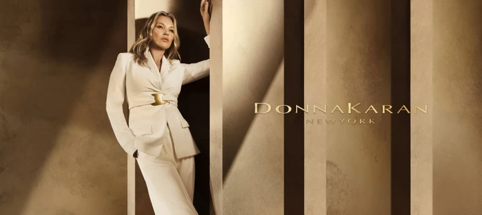 Kate Moss leaning against a concrete pillar in a white pantsuit for the Donna Karan Spring 2025 ad campaign.