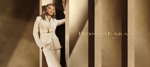Kate Moss leaning against a concrete pillar in a white pantsuit for the Donna Karan Spring 2025 ad campaign.