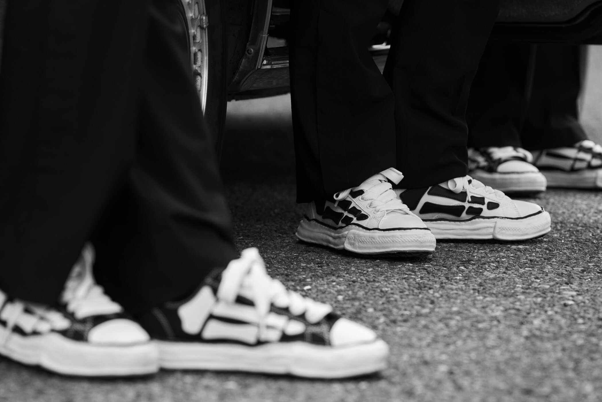 Close-up black and white shot of multiple people wearing dark trousers and chunky sneakers standing on a paved street.