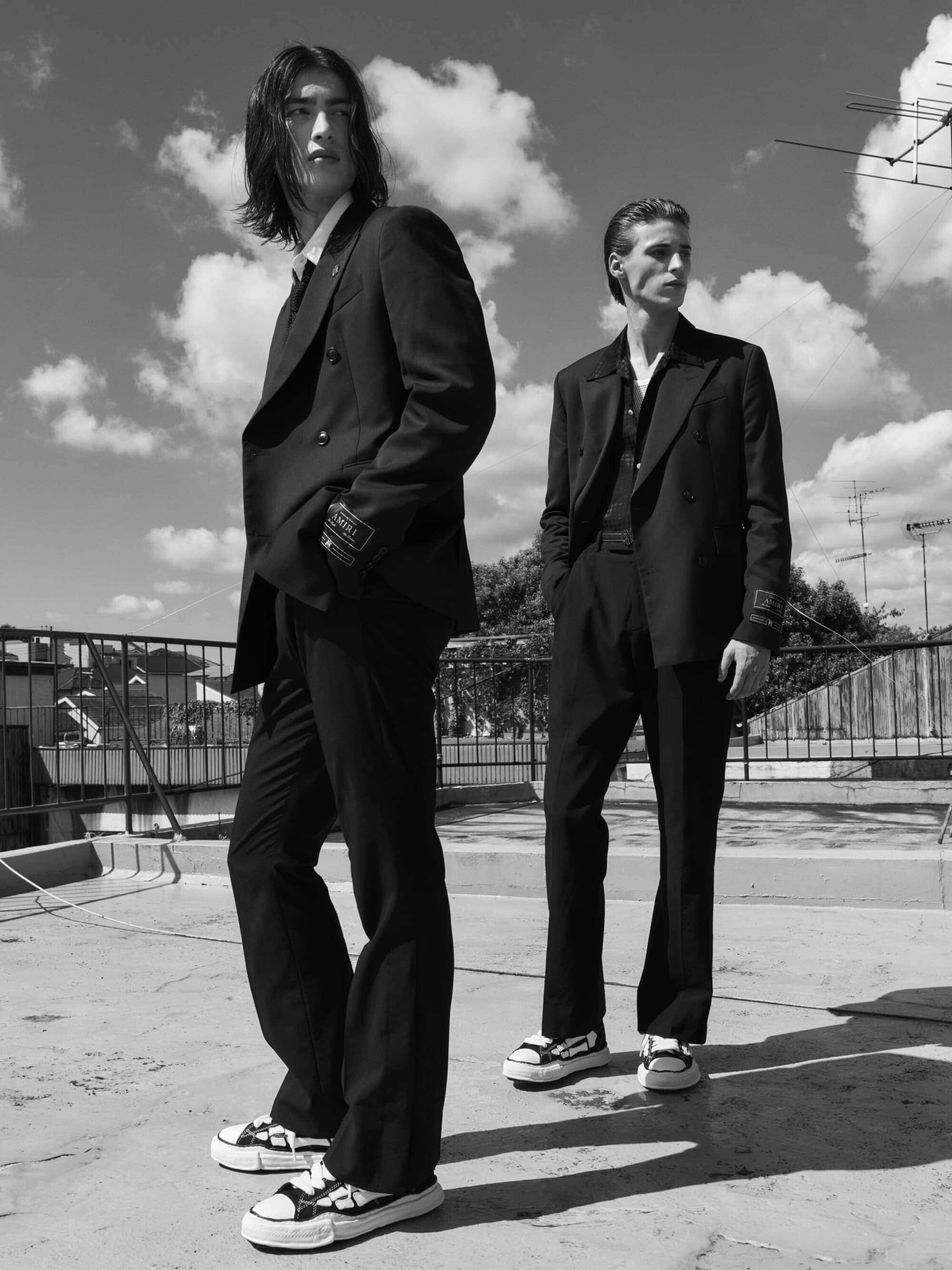 Two male models in dark suits and sneakers standing on a rooftop under a partly cloudy sky for an ad campaign.