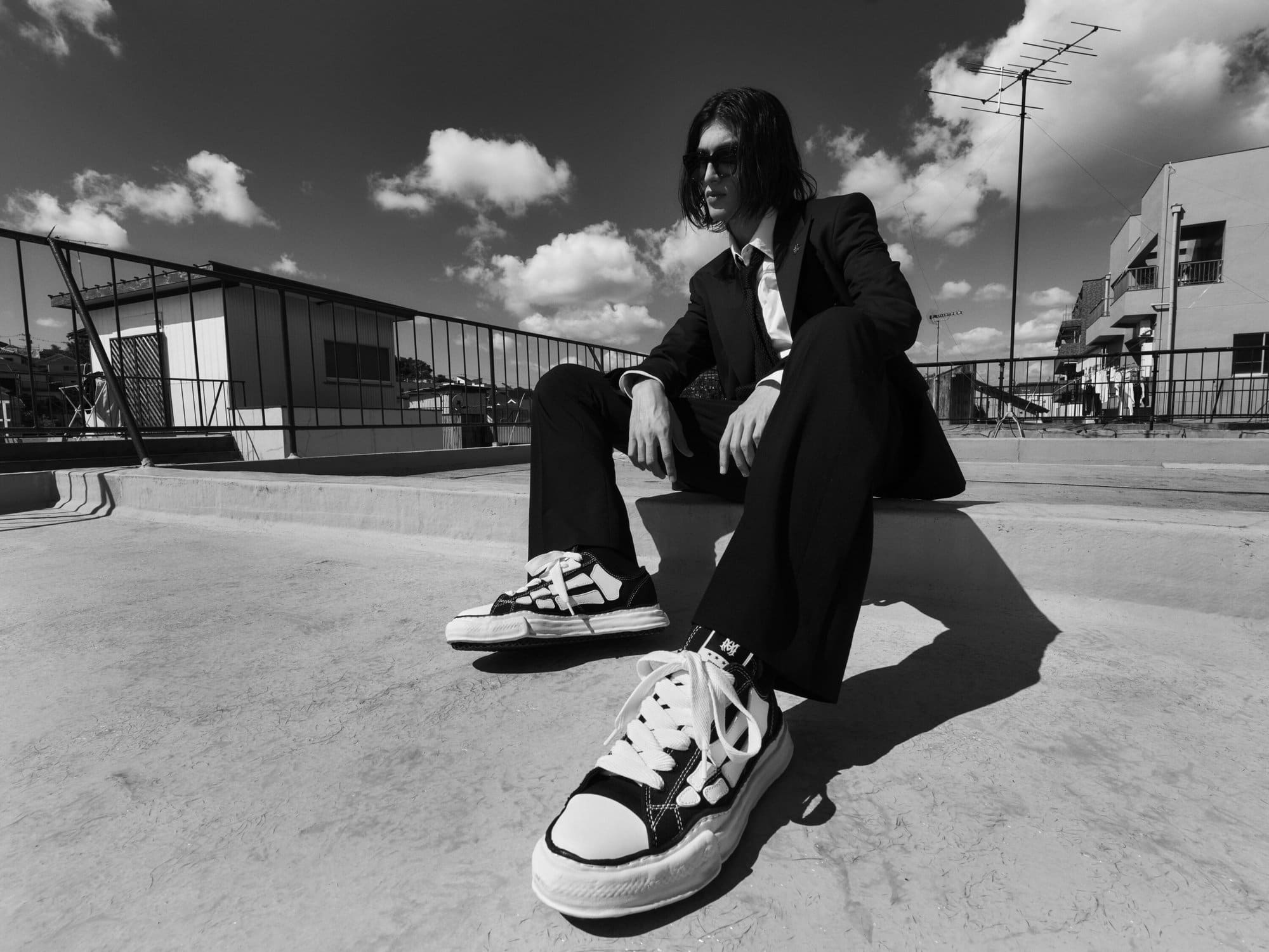 Man wearing dark suit and chunky sneakers seated on a rooftop under a partly cloudy sky.