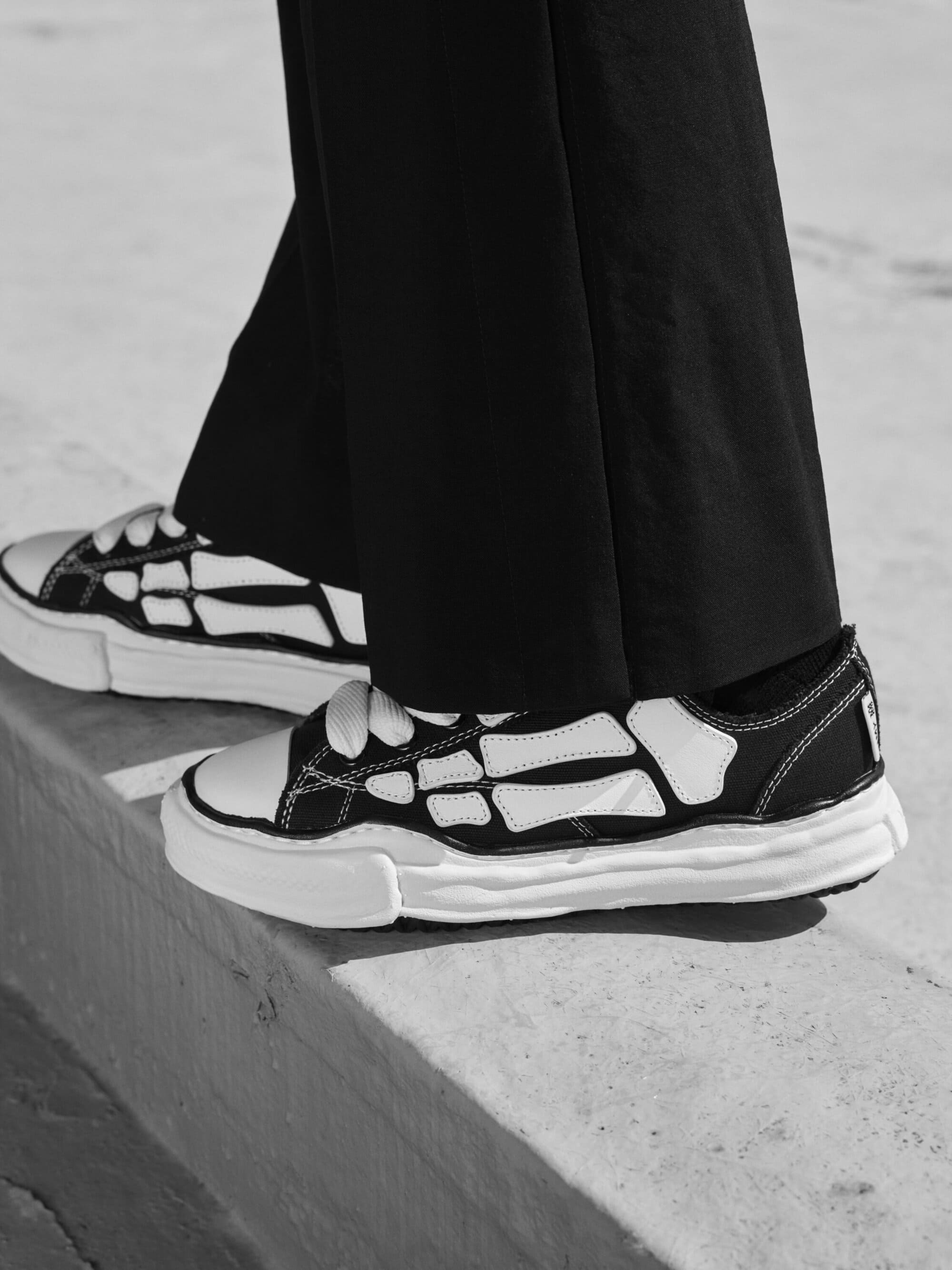 Close-up of black and white sneakers with chunky distressed soles and skeleton bone appliques worn with dark trousers