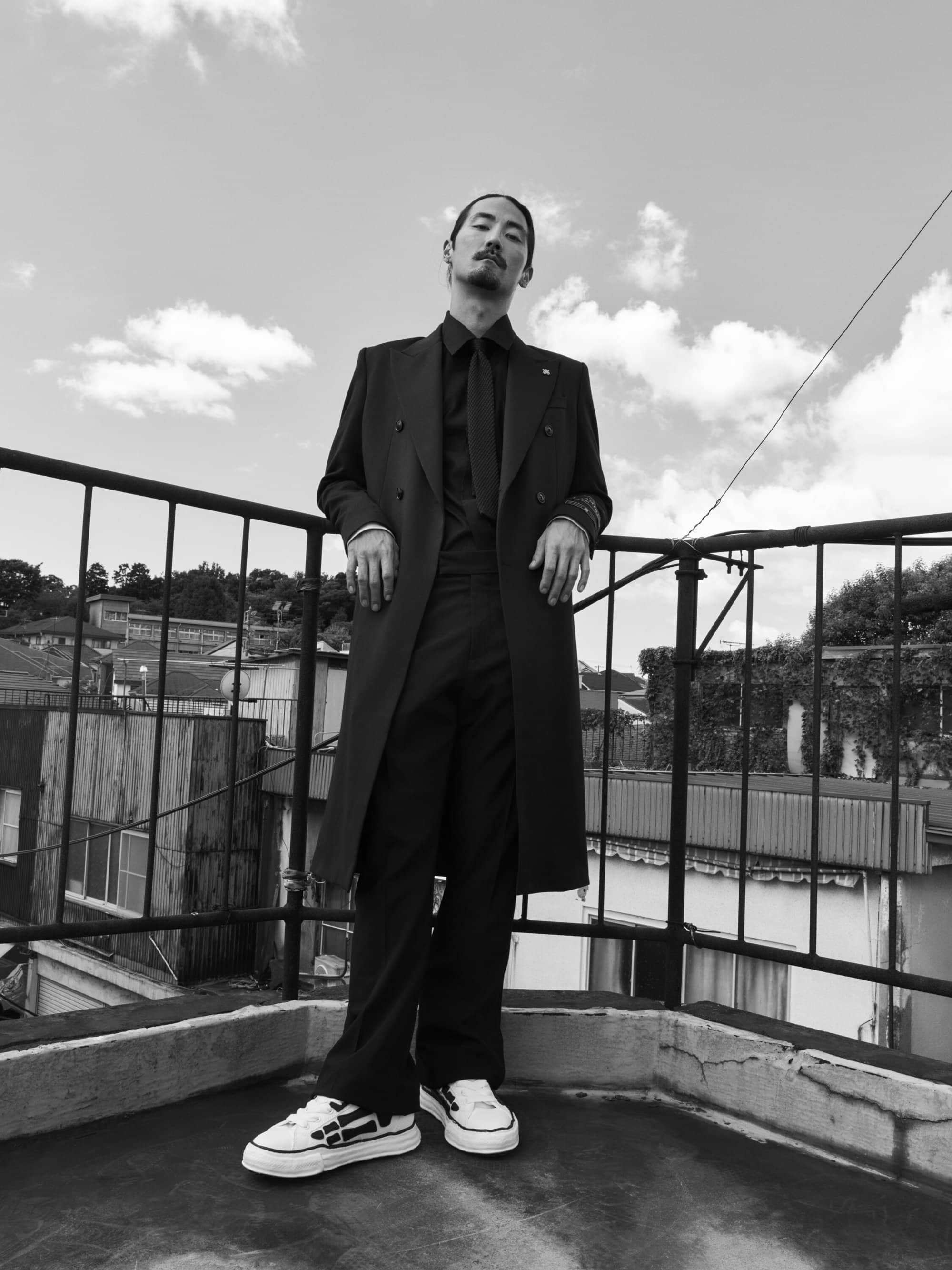 Man wearing a long dark coat and tie standing on a rooftop against a cloudy sky in a black and white fashion image.