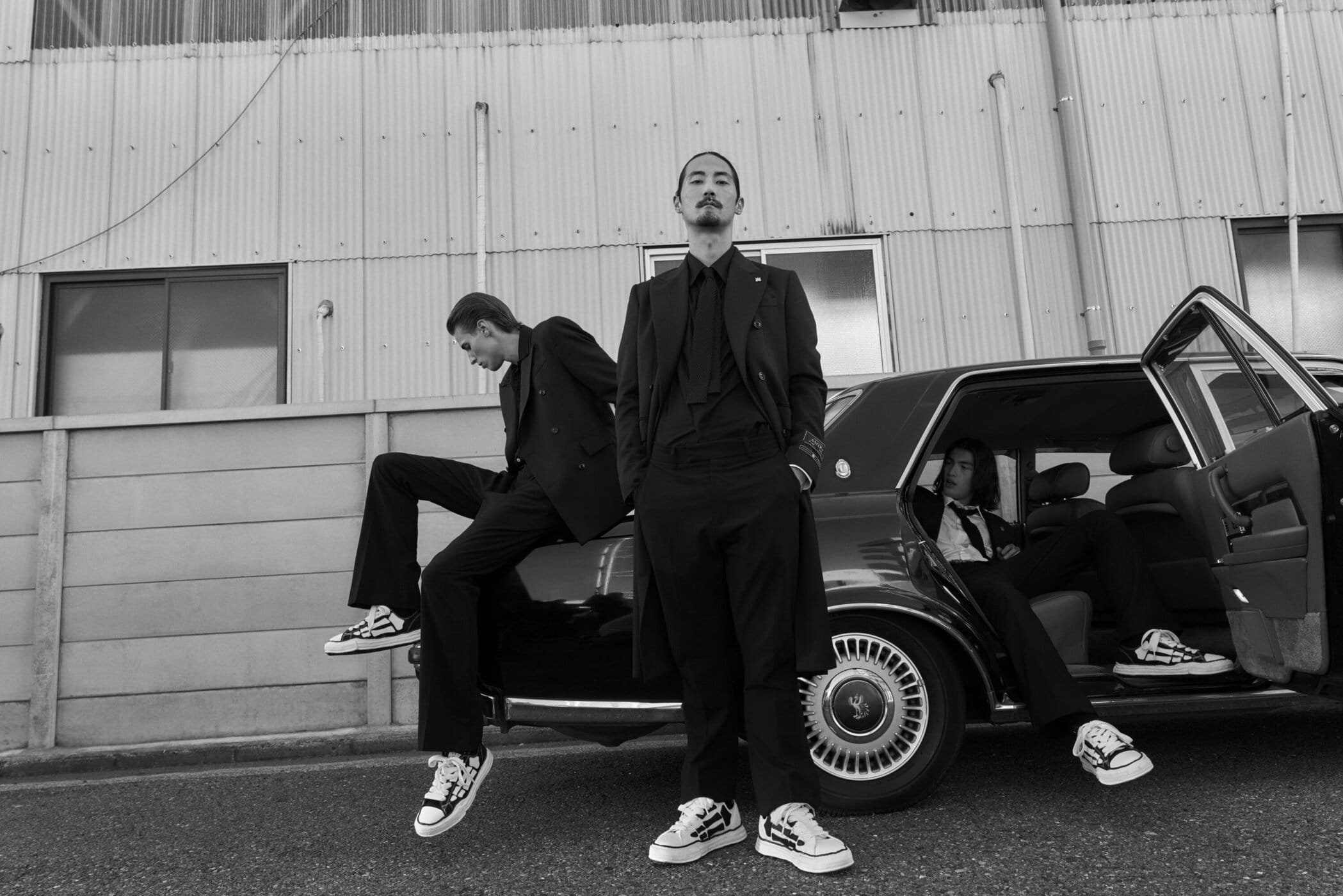 Three male models in dark suits and sneakers posing around a vintage car against an industrial building exterior