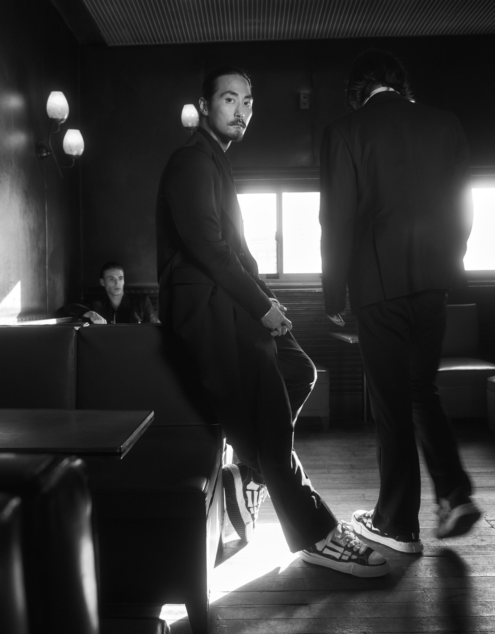 Man wearing dark jacket and sneakers leaning against a booth in a dark bar interior with another figure standing nearby