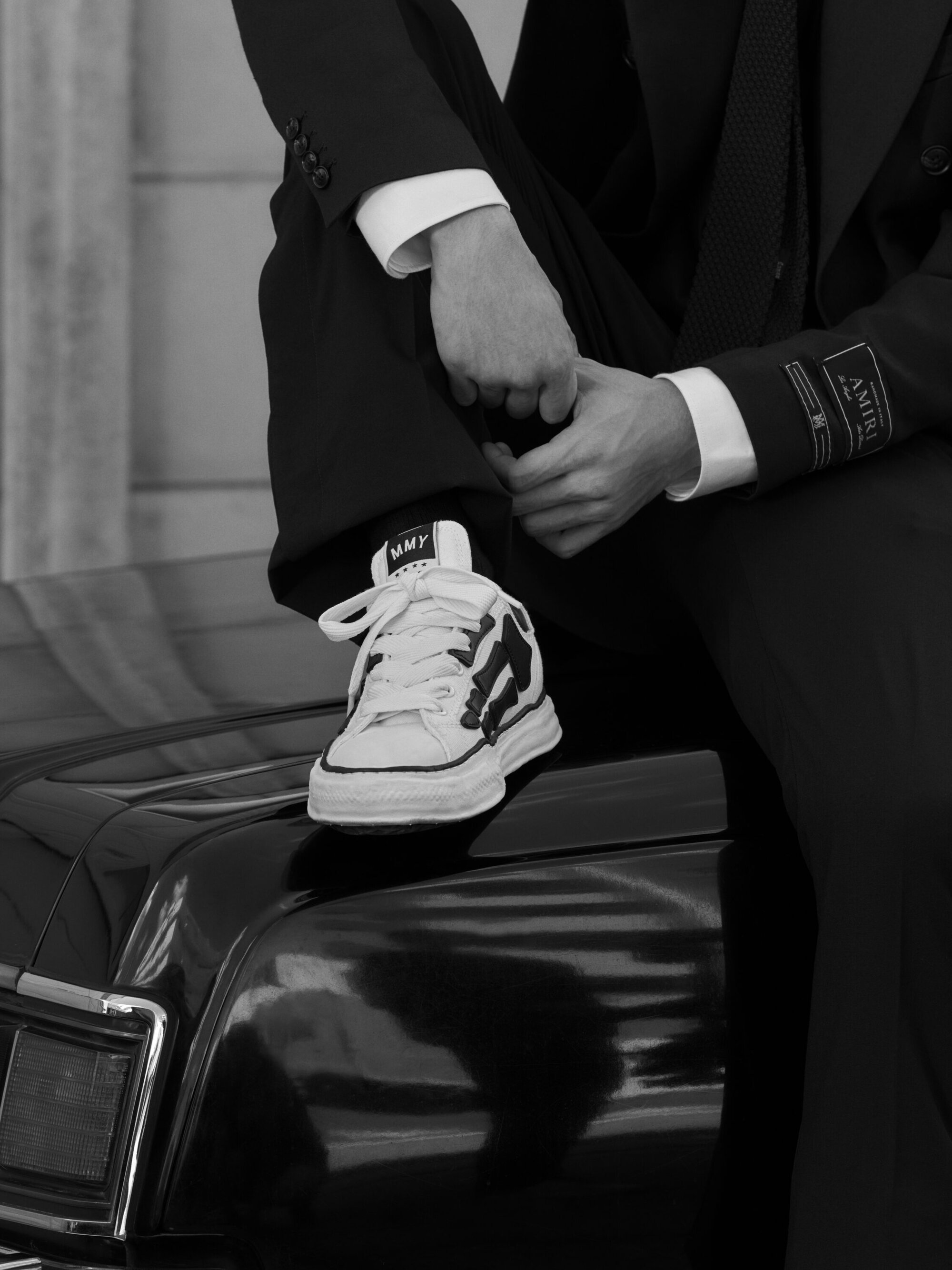 Close-up of a person in a black suit sitting on a car hood, wearing chunky sneakers.