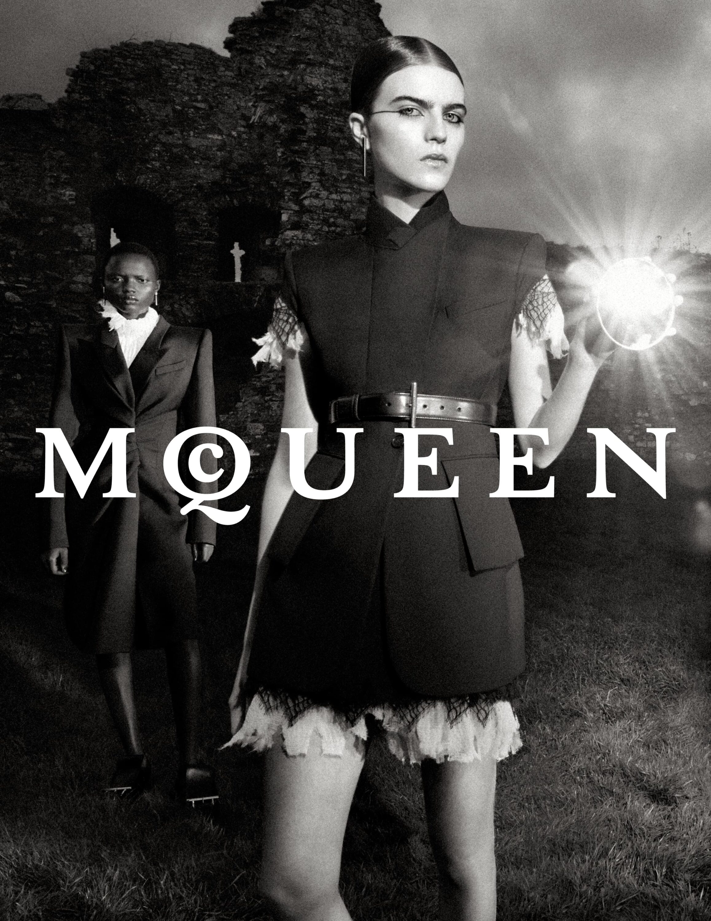 Black and white fashion campaign image featuring two models standing outdoors before a ruined stone building holding a glowing light source.