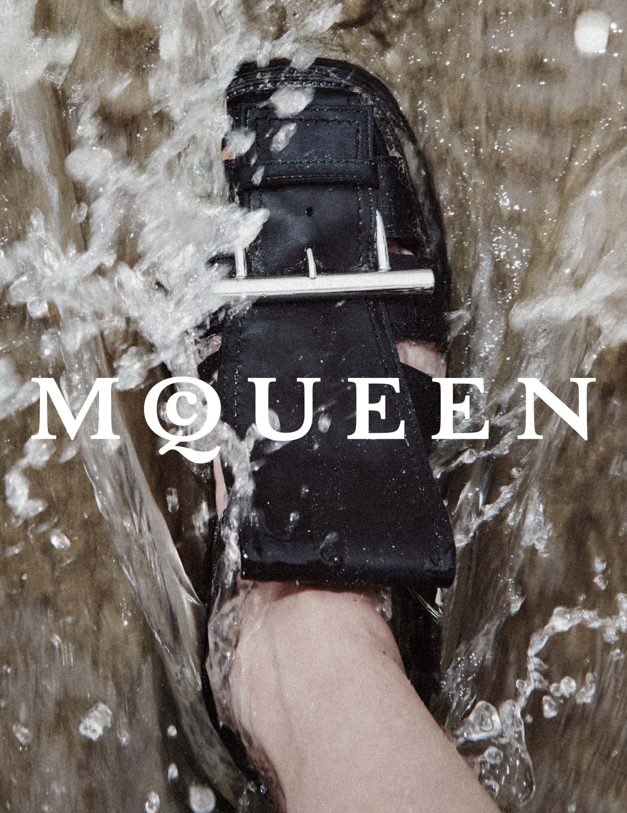 Close-up of a black buckled sandal submerged in splashing water with white lettering overlaid for McQueen Spring 2025 ad campaign