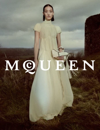 Model in a cream pleated dress holding a white handbag standing outdoors on a grassy hillside under a cloudy sky.