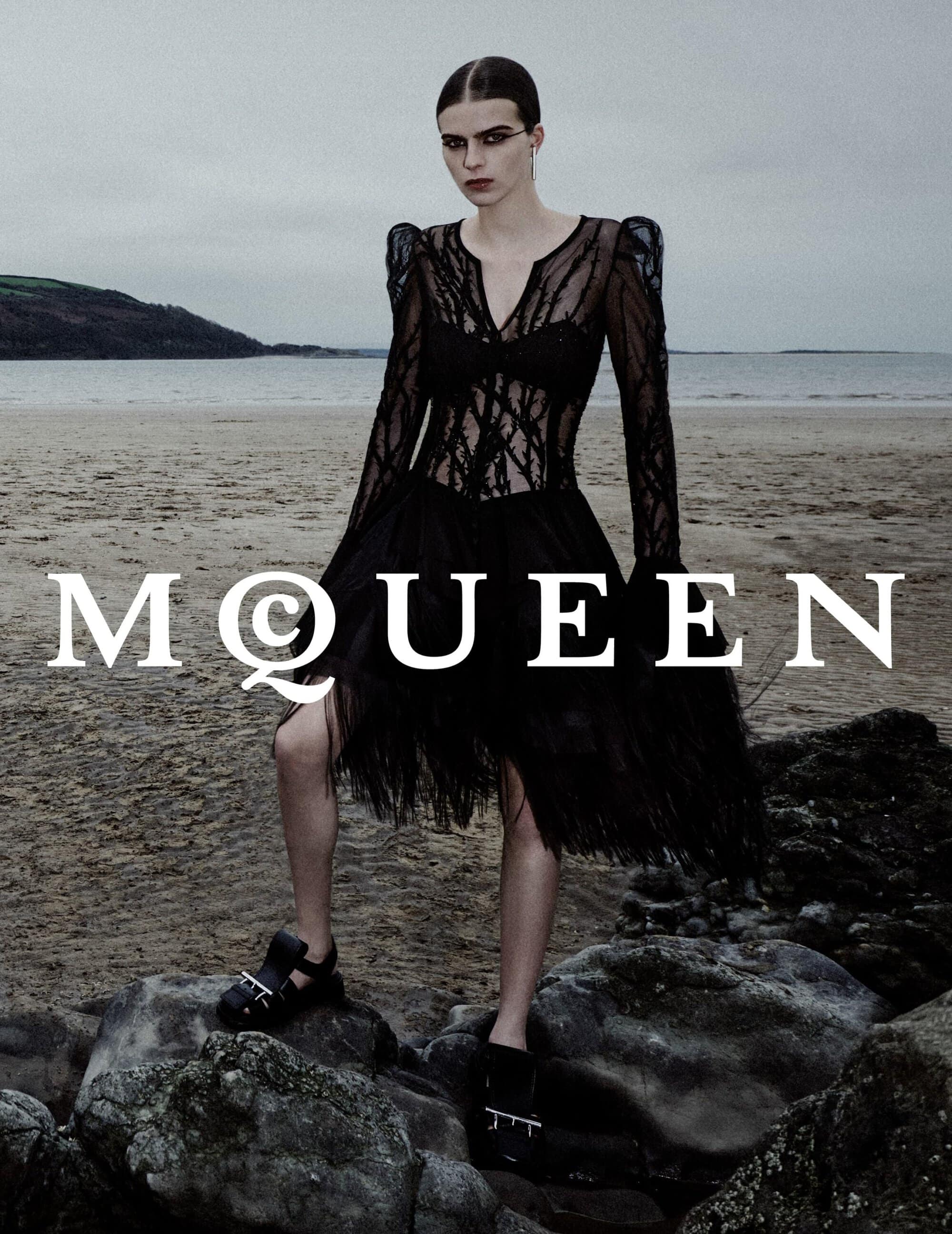 Model wearing a black lace dress standing on rocks near a coastal beach under an overcast sky for McQueen Spring 2025 campaign.