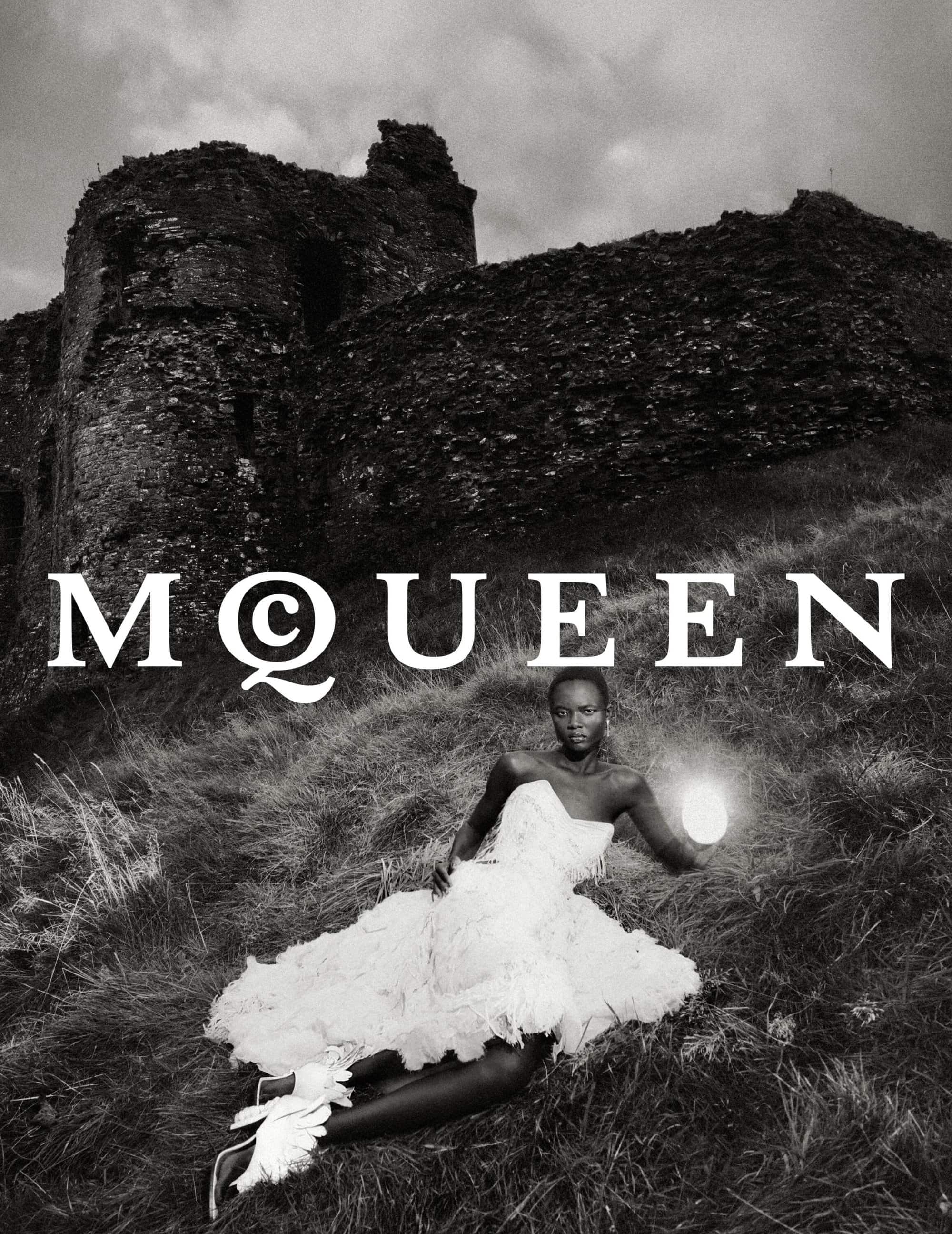 Model in a white ruffled dress lying on grassy hillside before a stone castle ruin, holding a glowing light source.