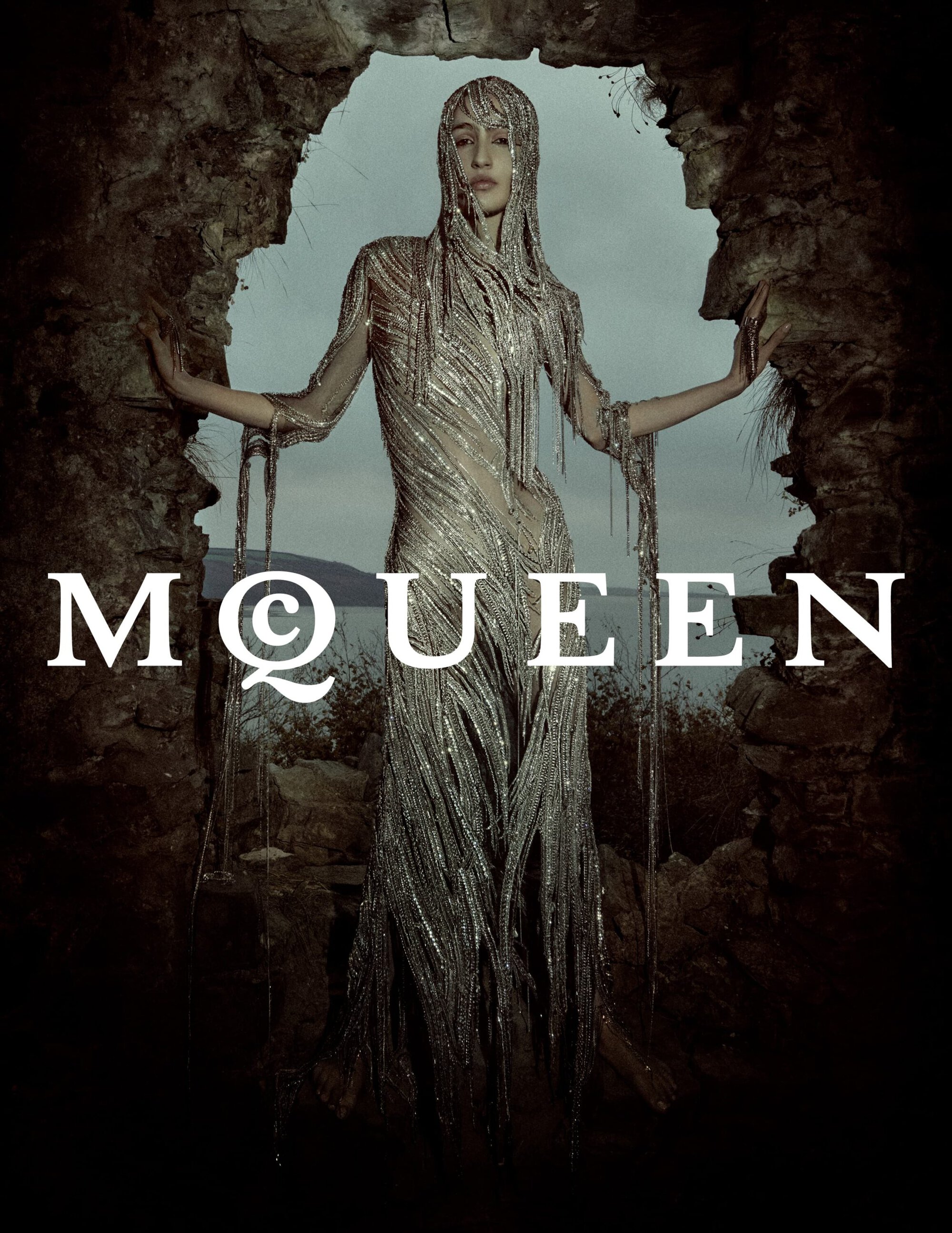 Model in a shimmering fringe dress standing inside a rough stone ruin opening for McQueen ad campaign.