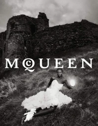 Model in a white ruffled dress lying on grassy hillside before a stone castle ruin, holding a glowing light source.